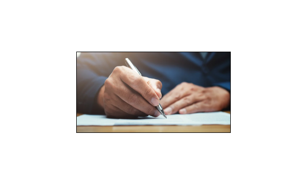 close up photo of two hands - one holding a silver pen and writing something on some blue paper atop a wooden desk